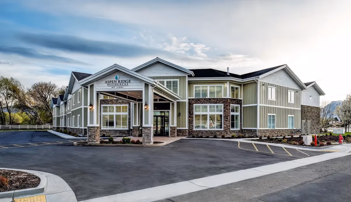 Front exterior of a two-story senior living building with a covered entrance, large windows, and adjacent parking area.