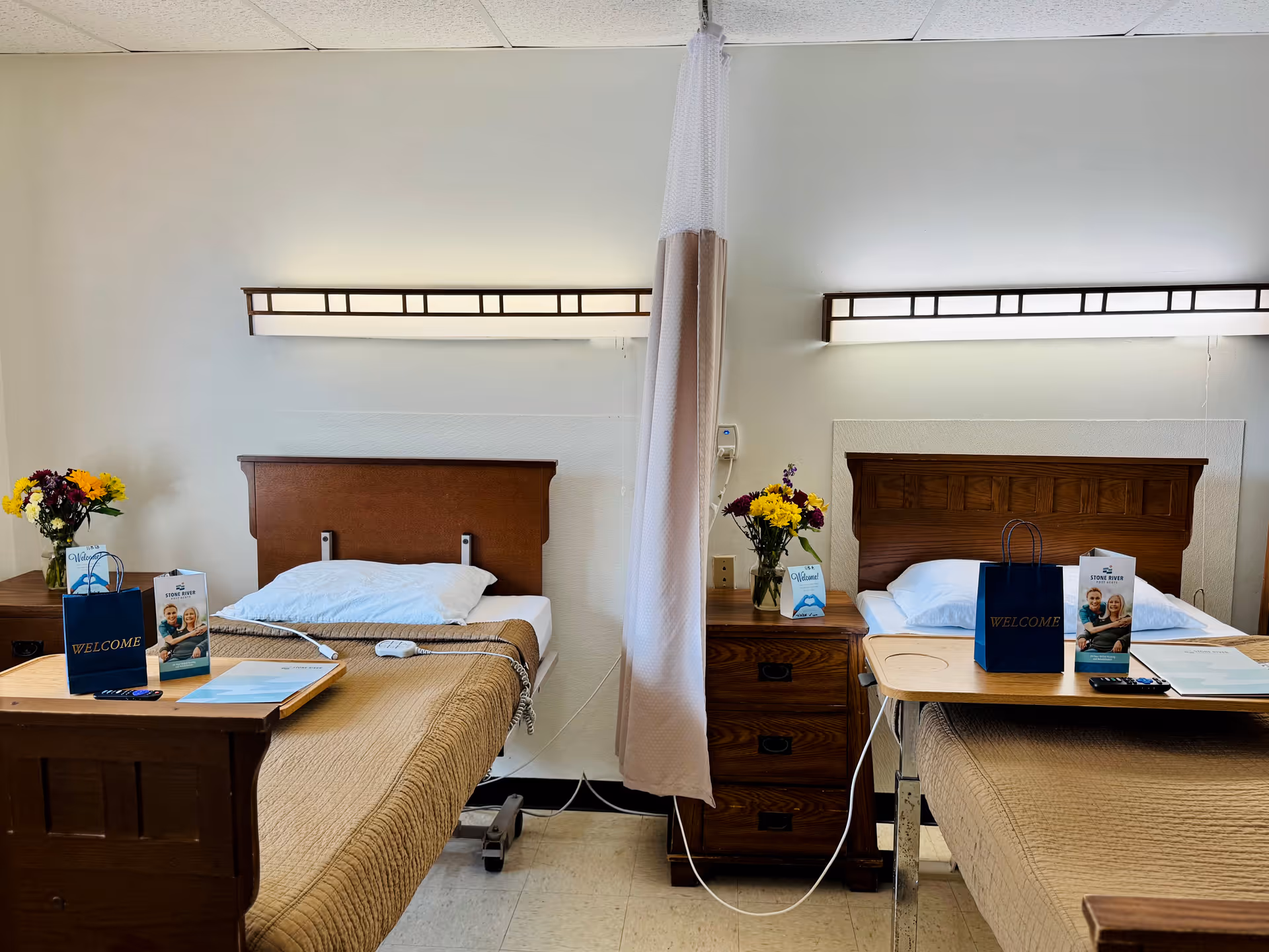 A shared hospital-style room with two beds separated by a privacy curtain. Each bed has a wooden headboard, a beige blanket, and a white pillow. Between the beds is a wooden nightstand with a vase of colorful flowers and welcome brochures. Overhead lights are mounted on the wall above each bed. Both beds have overbed tables with welcome bags and informational pamphlets.
