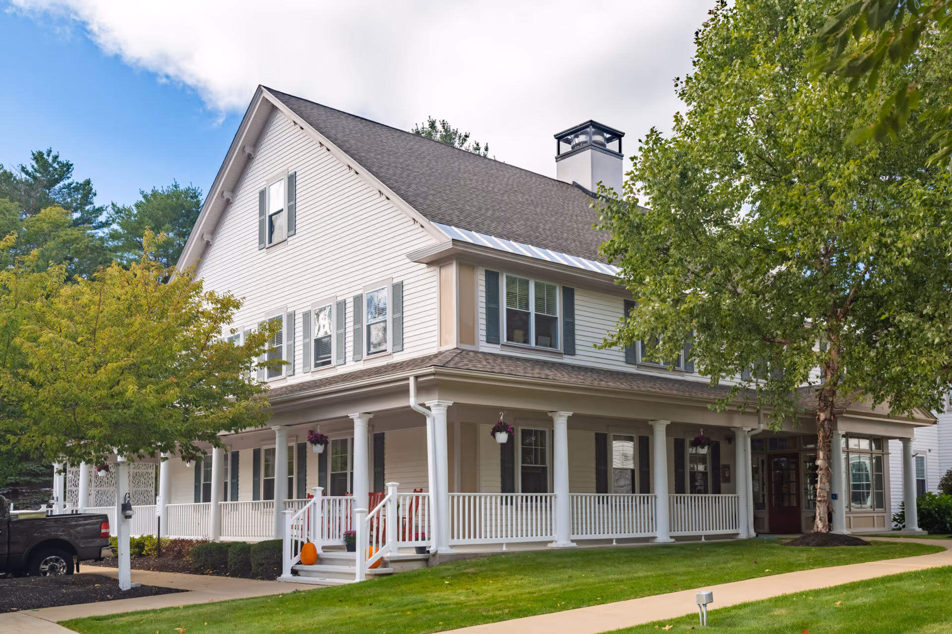 Exterior view of a large two-story white building with a wraparound porch, white columns, and green shutters. The building is surrounded by green grass, trees, and a sidewalk leading to the entrance. A black pickup truck is parked on the left side near the building.