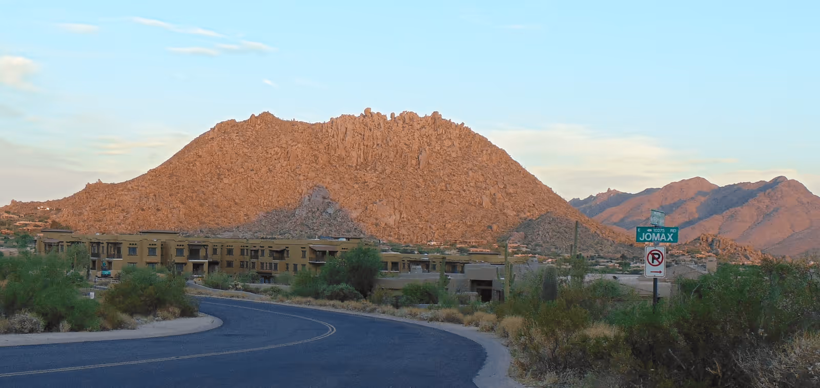 A winding road leading to a low-rise building complex with desert vegetation and large rocky mountains in the background under a partly cloudy sky.