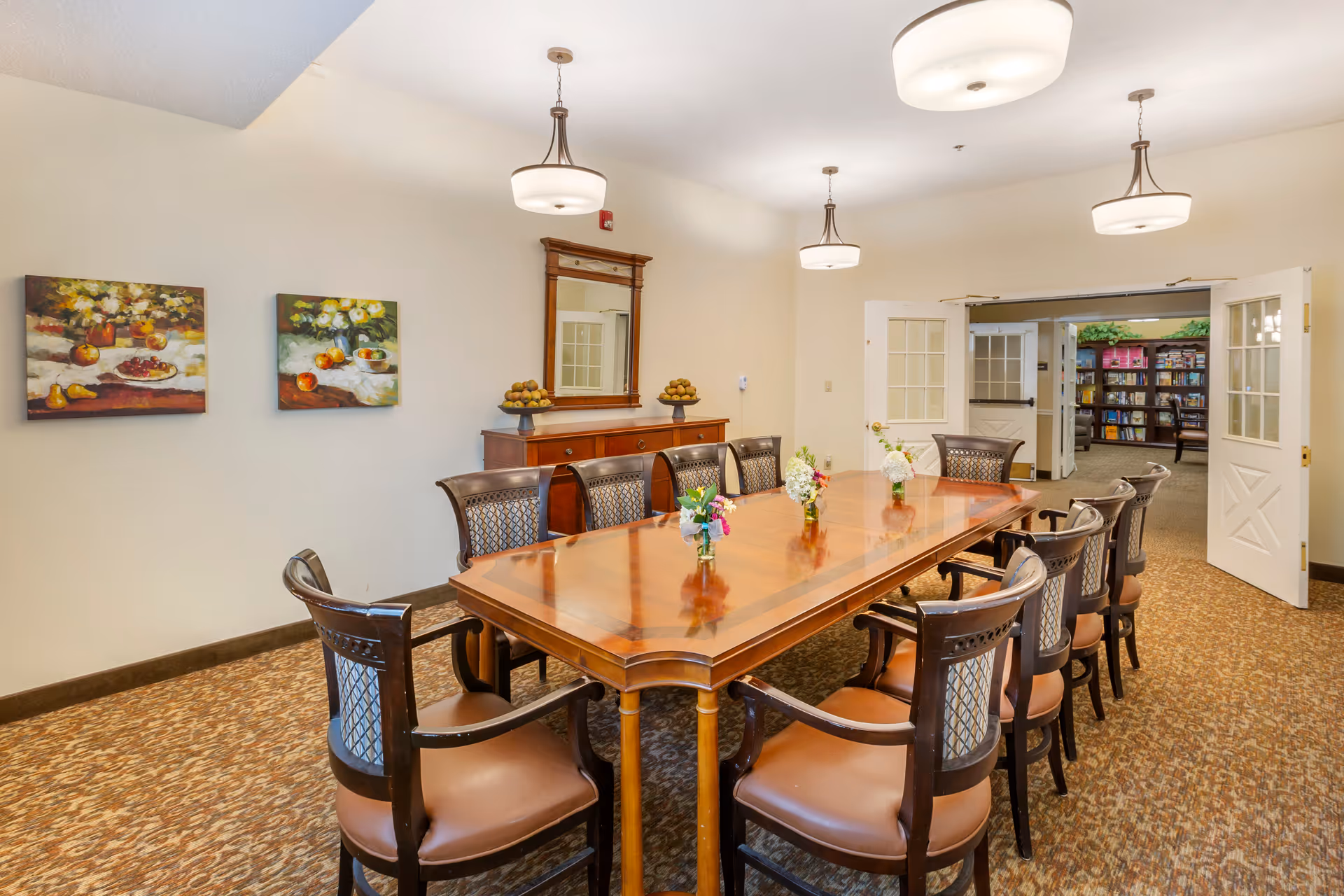 A well-lit dining room with a long wooden table surrounded by ten chairs. The table has small flower arrangements placed along its center. The room features beige walls, two paintings of fruit on the left wall, a wooden sideboard with a mirror above it, and double doors leading to a room with bookshelves.