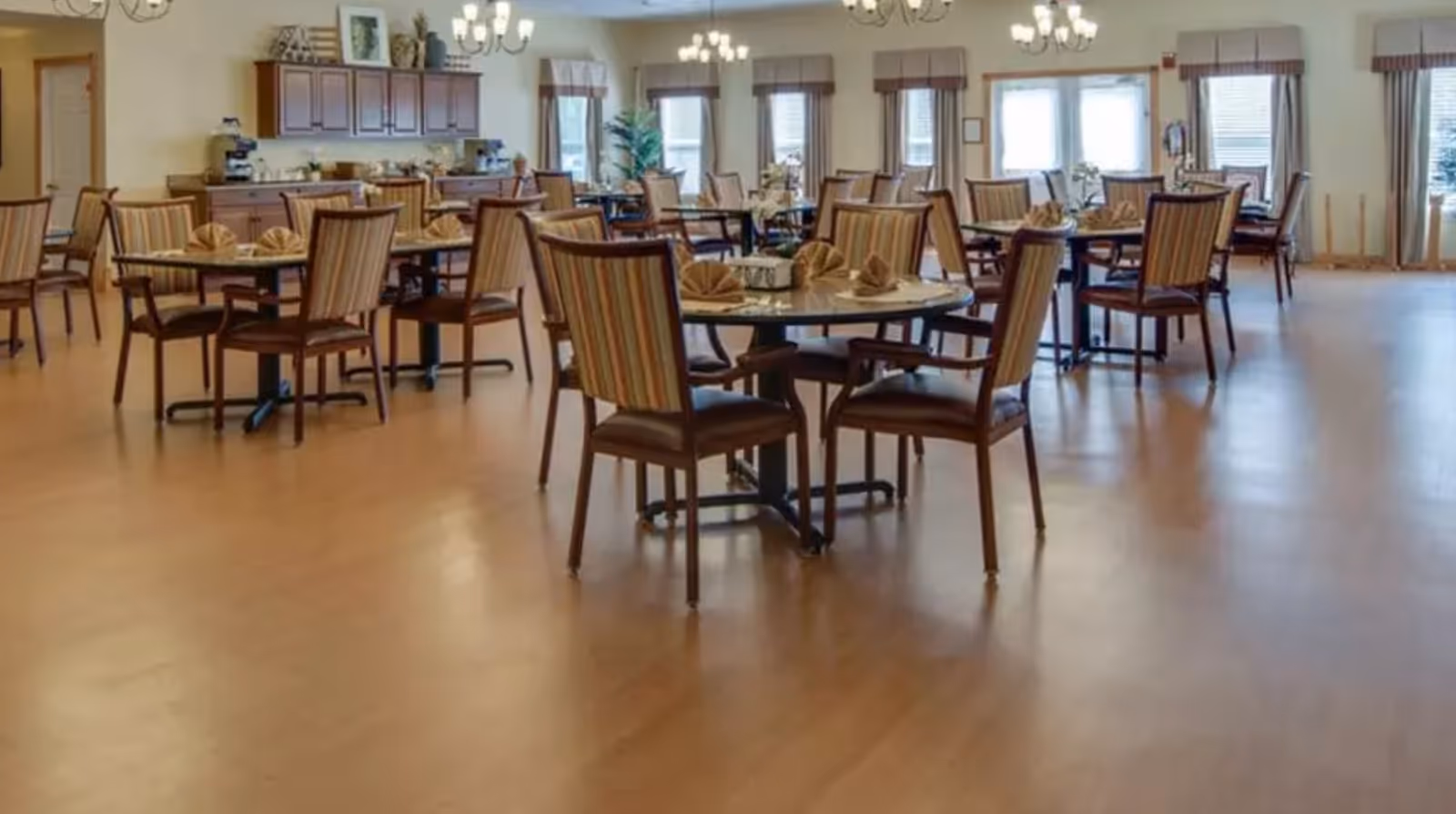 A spacious dining room with multiple round tables and striped cushioned chairs. Each table is set with folded napkins and a centerpiece. The room has large windows with curtains, wooden flooring, and chandeliers hanging from the ceiling. There is a wooden cabinet with decorative items and a coffee machine along one wall.