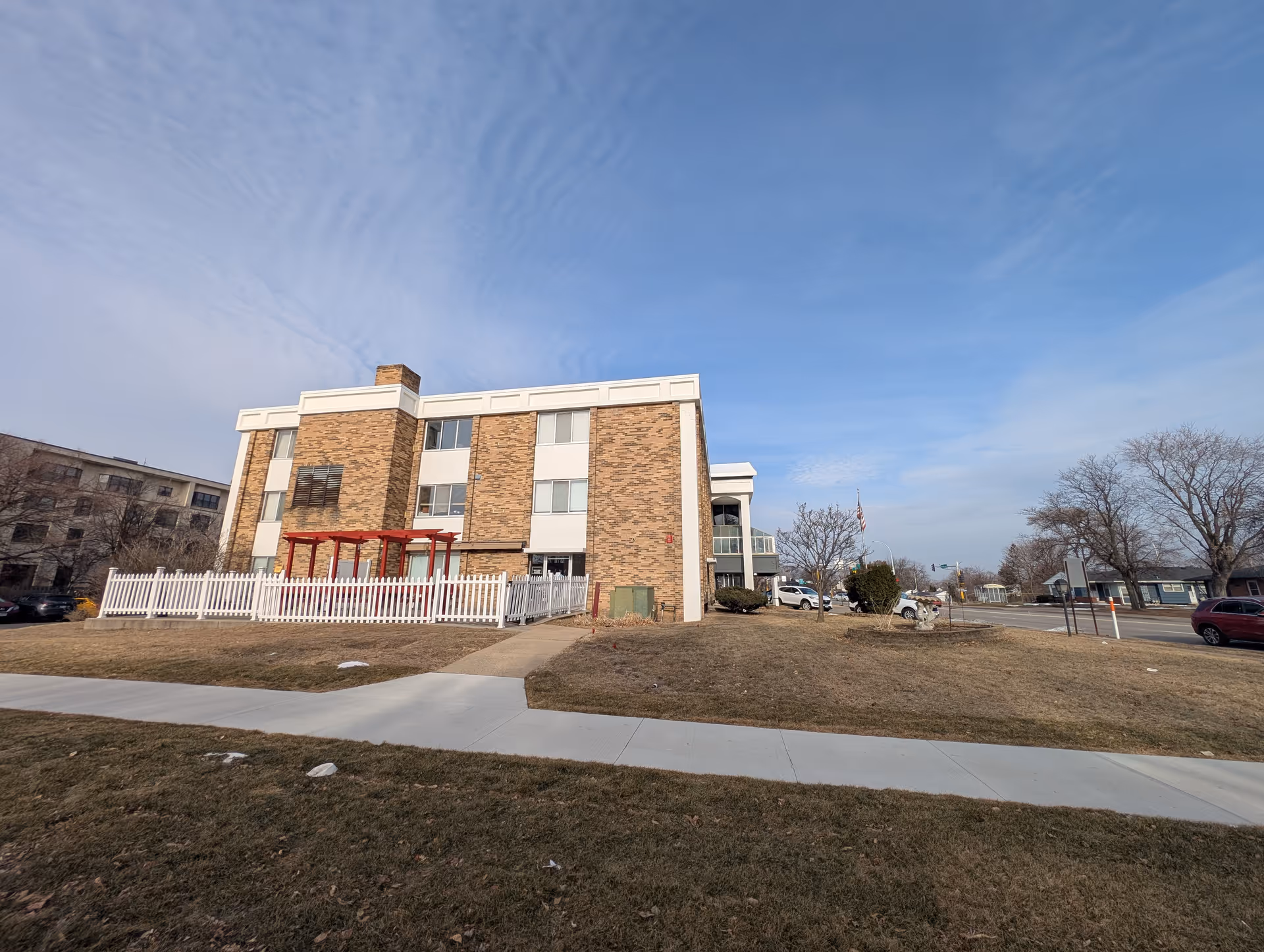 Exterior view of a three-story brick building with white trim, a white picket fence enclosing a small patio area with a red pergola, and a sidewalk leading up to the building. The sky is partly cloudy and there are some leafless trees and parked cars nearby.