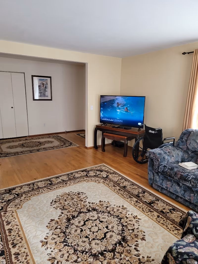 A living room with beige walls and wooden flooring. There is a patterned area rug on the floor, a patterned sofa on the right, and a wheelchair next to it. A flat-screen TV is placed on a wooden table against the wall, displaying an image of people swimming. A framed picture hangs on the wall near a closed closet with double doors.