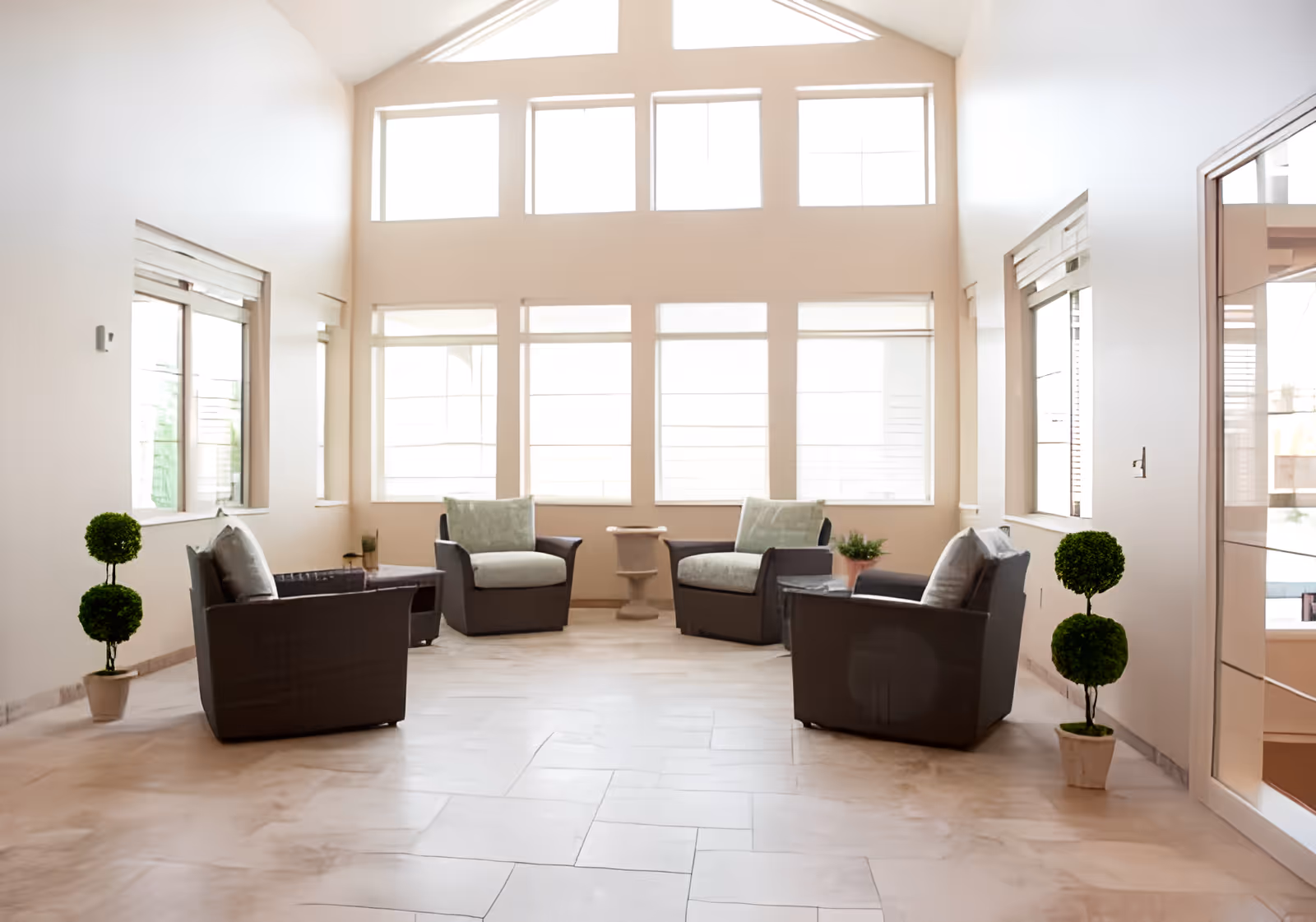 Bright and spacious sitting area with four cushioned armchairs arranged around two small tables, large windows letting in natural light, and two potted topiary plants on either side.