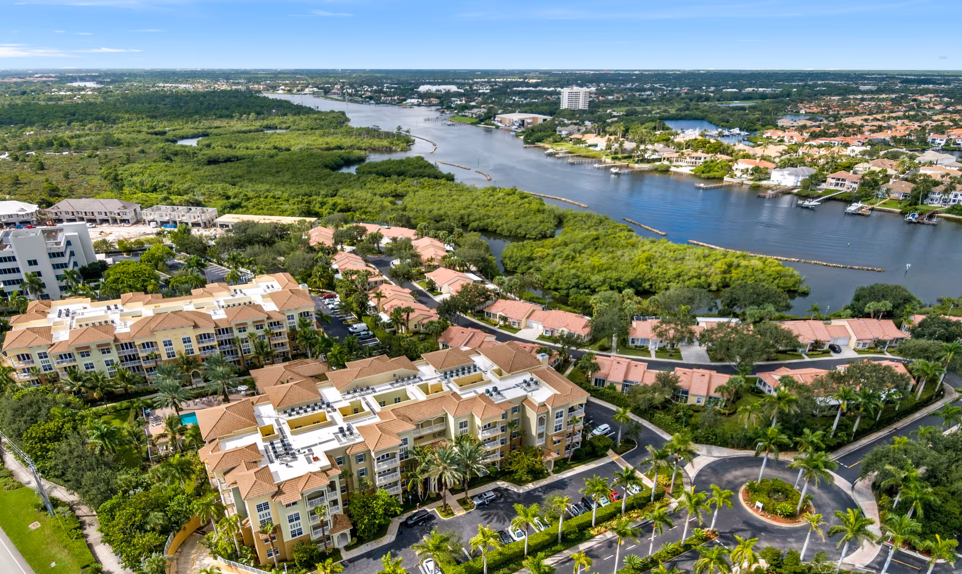 Aerial view of a waterfront senior living complex with multiple Mediterranean-style buildings, landscaping, roads, and a river.