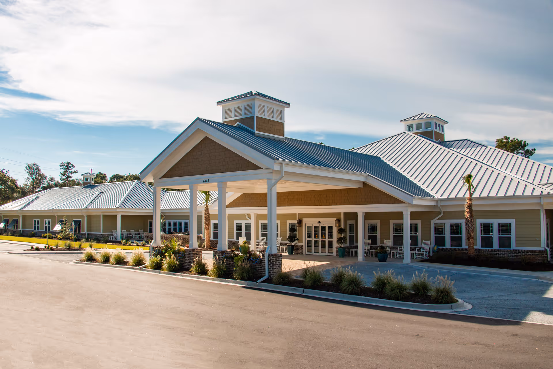 Exterior view of Arbor Landing at Pawleys, showing a single-story building with a metal roof, covered entrance with white pillars, and landscaping with small palm trees and shrubs under a partly cloudy sky.