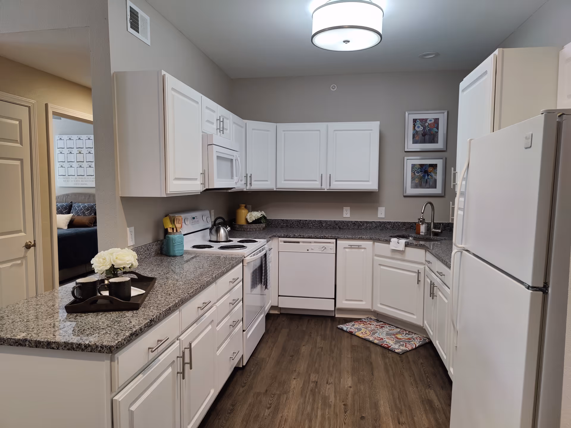 A modern kitchen with white cabinets, granite countertops, and wood flooring. The kitchen features a white refrigerator, stove with oven, microwave, and dishwasher. There are two framed floral artworks on the wall above the sink, and a colorful patterned mat on the floor in front of the sink. A tray with two black mugs and white flowers is placed on the countertop. A doorway to a bedroom is visible on the left side.