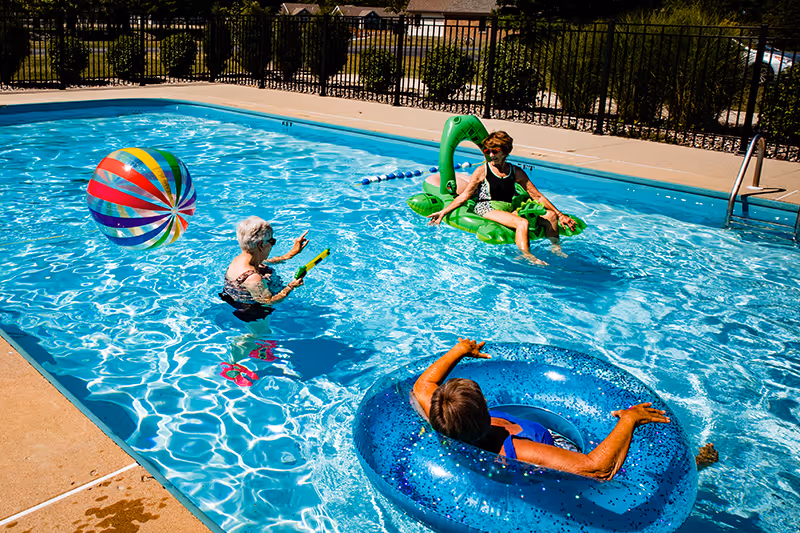 Three elderly women enjoying a sunny day in an outdoor swimming pool. One woman is floating on a green inflatable shaped like a dinosaur, another is sitting in a blue inflatable ring, and the third is standing in the water holding a water toy. A colorful beach ball is floating nearby, and a black metal fence surrounds the pool area with greenery and houses visible in the background.