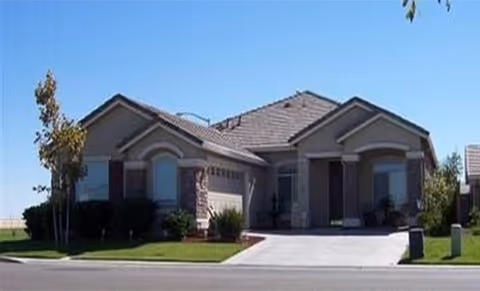 Single-story residential building with a tiled roof, two garage doors, and a driveway. The house is surrounded by a well-maintained lawn with a few small trees and shrubs. The sky is clear and blue.