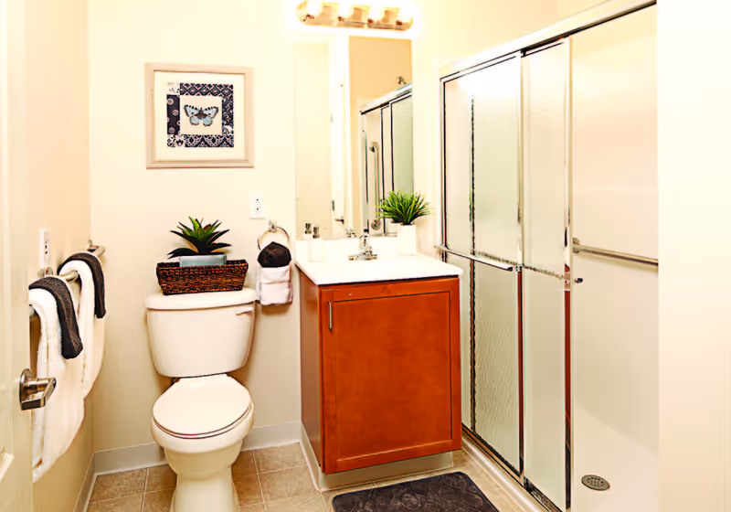A clean and bright bathroom featuring a toilet with a small plant on top, a wooden vanity with a white countertop and a potted plant, a large mirror above the sink, a glass-enclosed shower with sliding doors, and two towel racks holding white and dark gray towels. A framed butterfly picture hangs on the wall above the toilet.