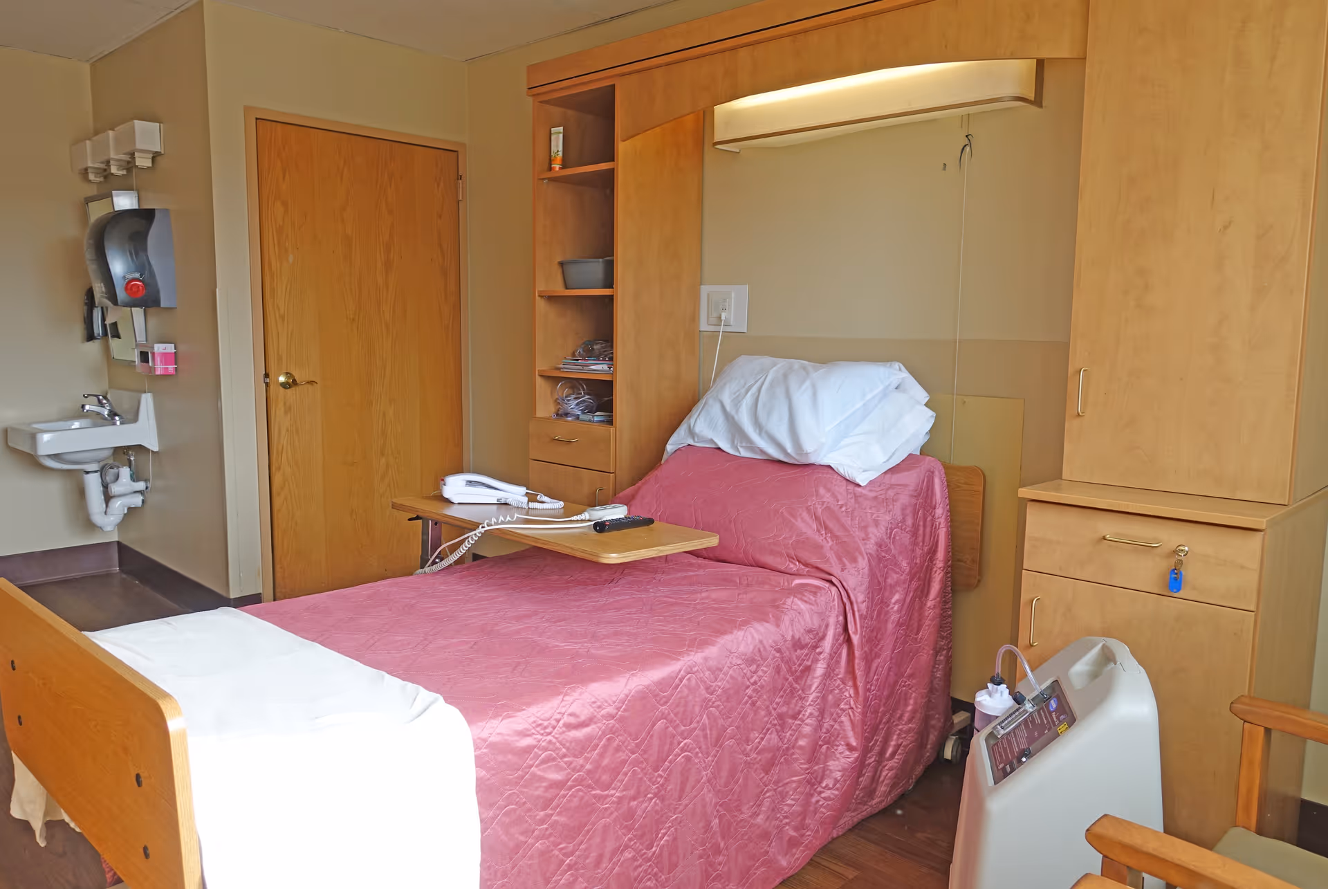 A nursing facility room with a single bed covered in a pink quilt, a white pillow, and a white towel at the foot of the bed. The bed has an overbed table with a telephone and remote control on it. There is a wooden cabinet with shelves and drawers behind the bed, a wooden door, a wall-mounted sink with soap dispensers, and a medical device on the floor next to the bed.