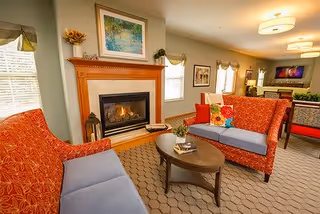 A cozy living room area with two red patterned sofas with blue cushions, a wooden coffee table with books and a small plant, a fireplace with a wooden mantel, and framed artwork above it. The room is well-lit with ceiling lights and natural light from windows with valances. In the background, there is a dining area with a table and chairs.