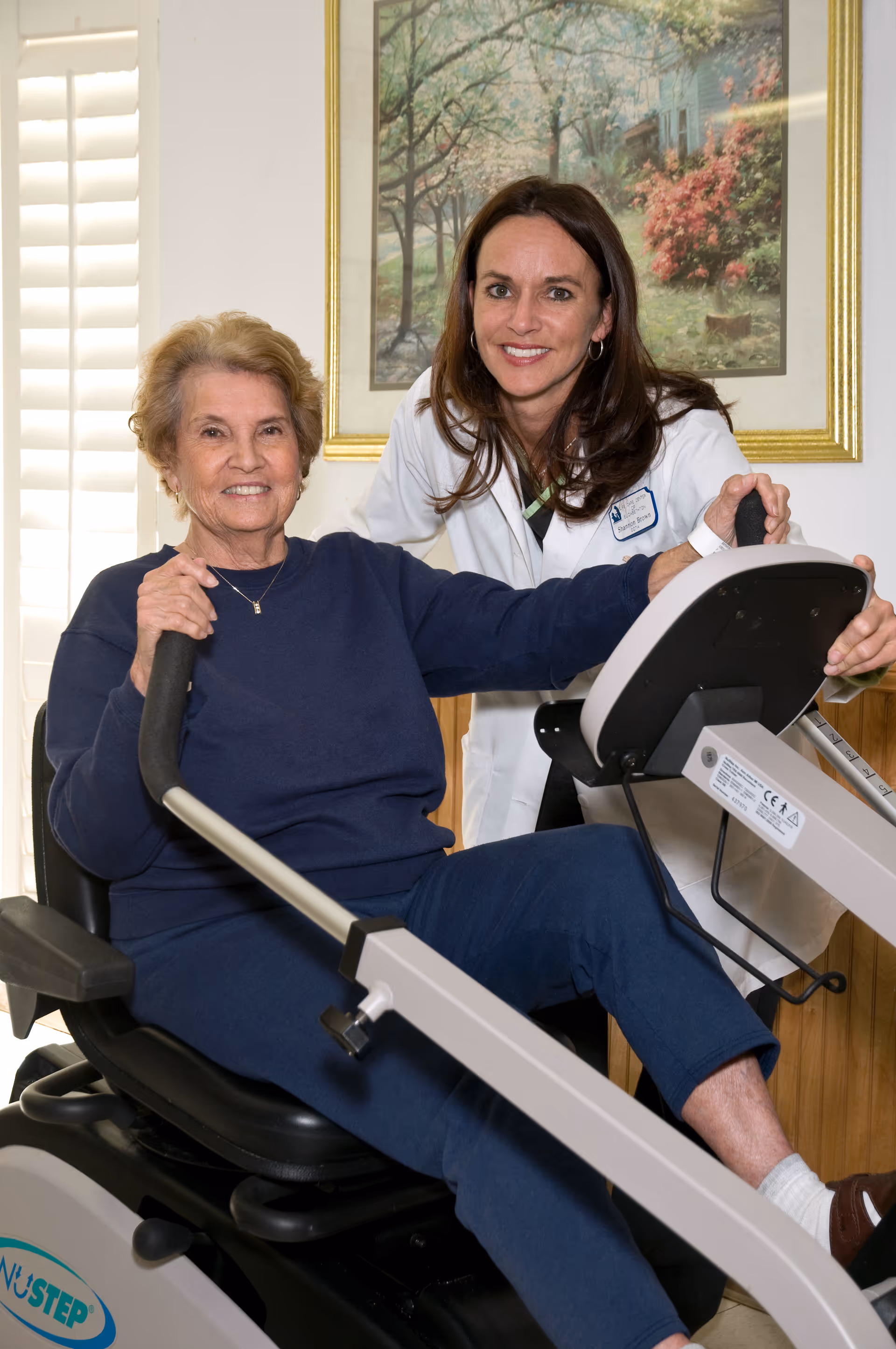 An elderly woman sitting on a NuStep exercise machine, smiling, with a female healthcare professional in a white coat standing beside her, also smiling. They are indoors with a framed painting and window shutters in the background.
