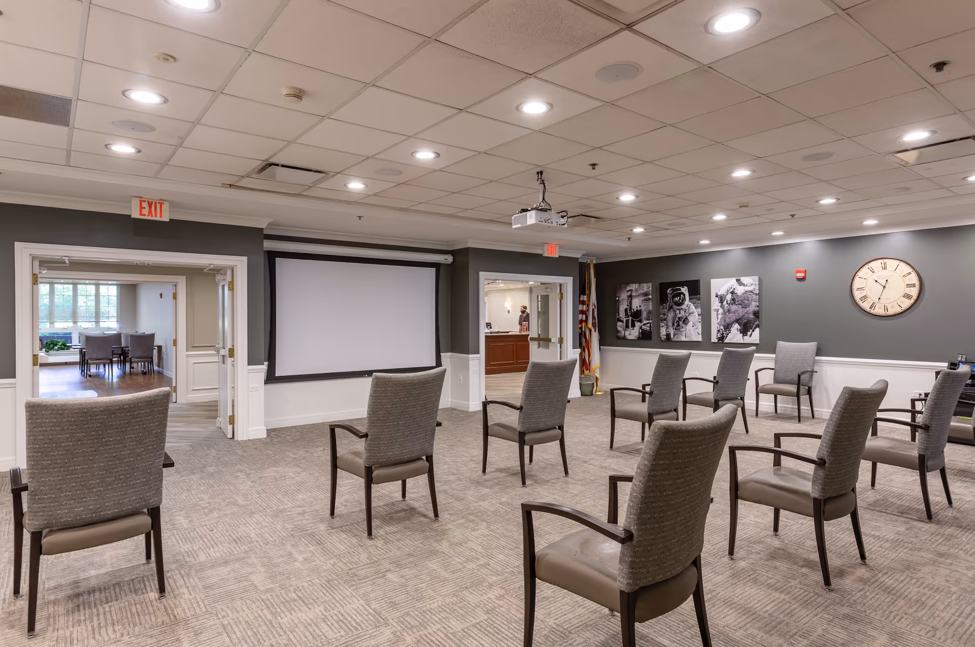 A meeting or presentation room with several chairs arranged in rows facing a large projection screen on the wall. The room has a carpeted floor, gray walls with white wainscoting, and a ceiling with recessed lighting. On one wall, there are three black and white framed pictures and a large clock. Two doorways lead to other rooms, one showing a dining area with tables and chairs, and the other showing a reception desk with a person behind it. An American flag is placed near the reception area.