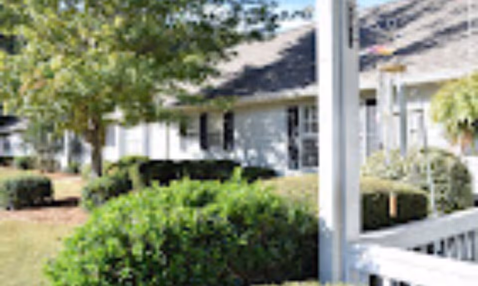 View of the exterior of a residential building with white siding, black shutters, and a shingled roof, surrounded by green bushes and trees on a sunny day.