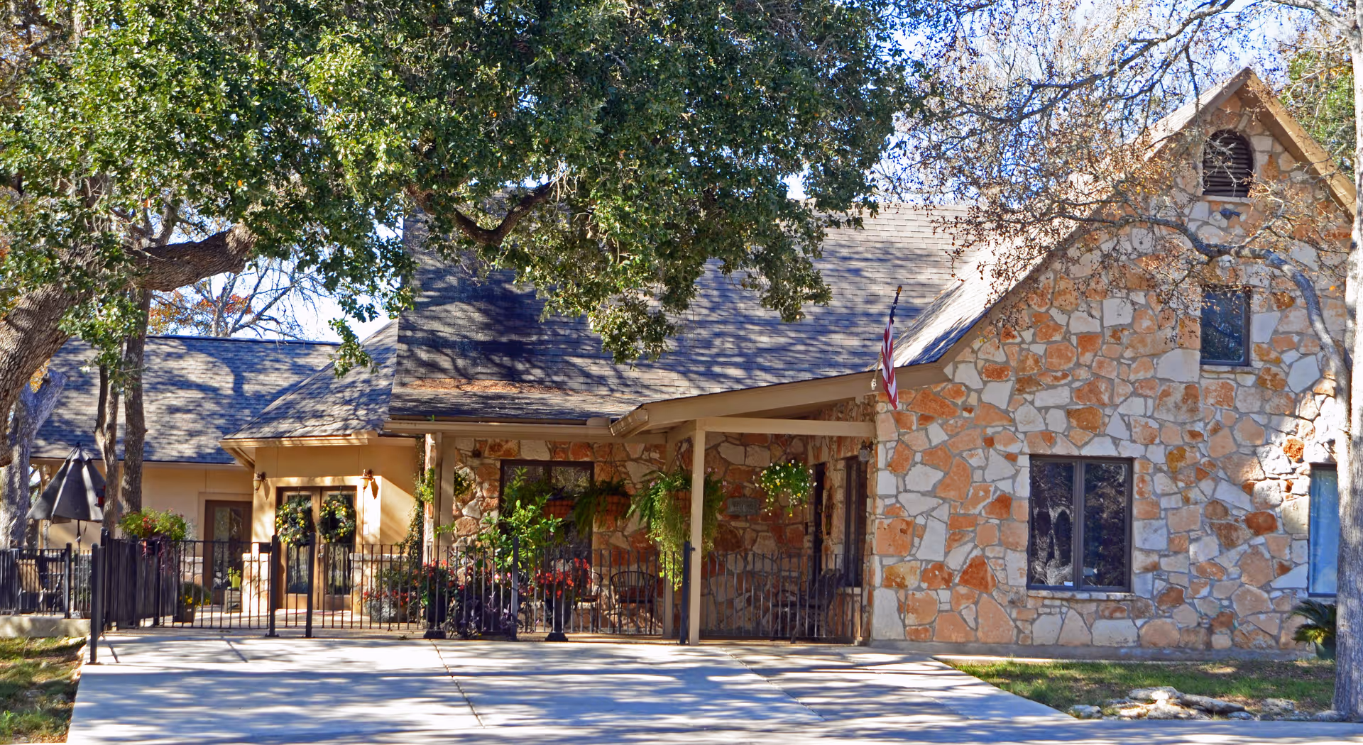 Exterior view of a stone and stucco building with a sloped roof, surrounded by trees and a paved driveway. The entrance is decorated with hanging plants and an American flag is displayed near the front porch.