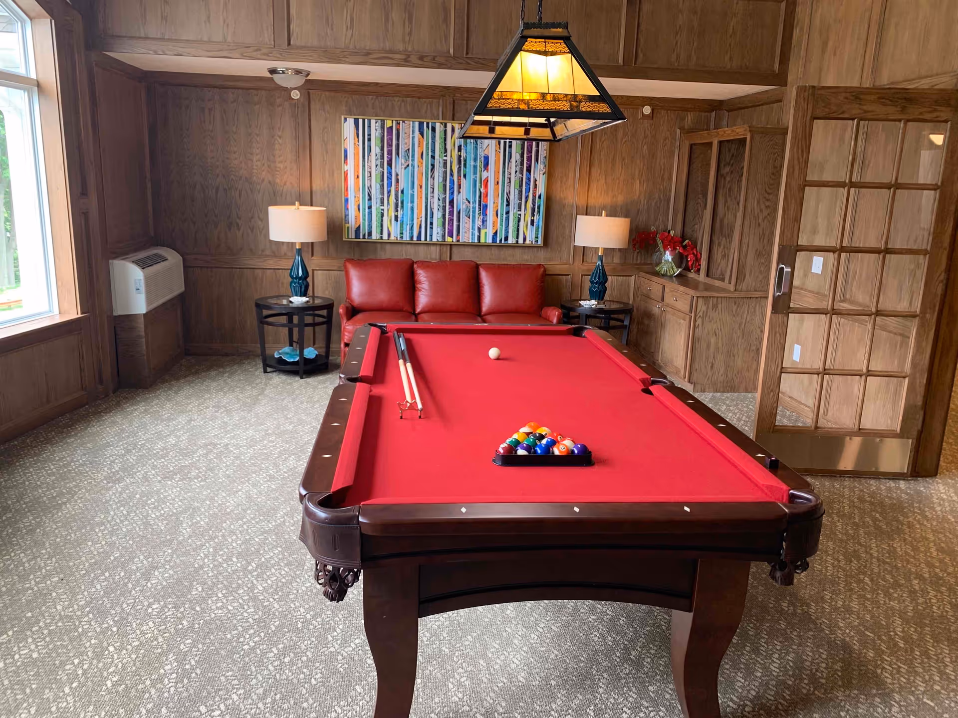 A cozy game room with a red felt pool table in the center, a red leather couch against a wood-paneled wall, two side tables with blue lamps, a colorful abstract painting above the couch, and a hanging light fixture above the pool table. There is a window on the left side and a wooden door with glass panels on the right.