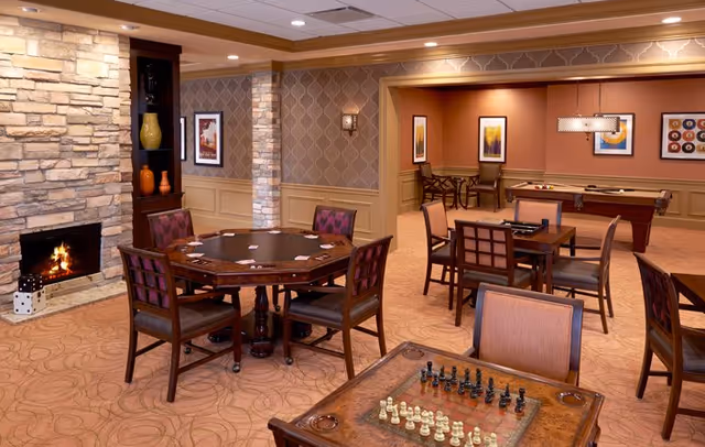 A warm senior living common room with card and chess tables, a pool table, fireplace, and seating.