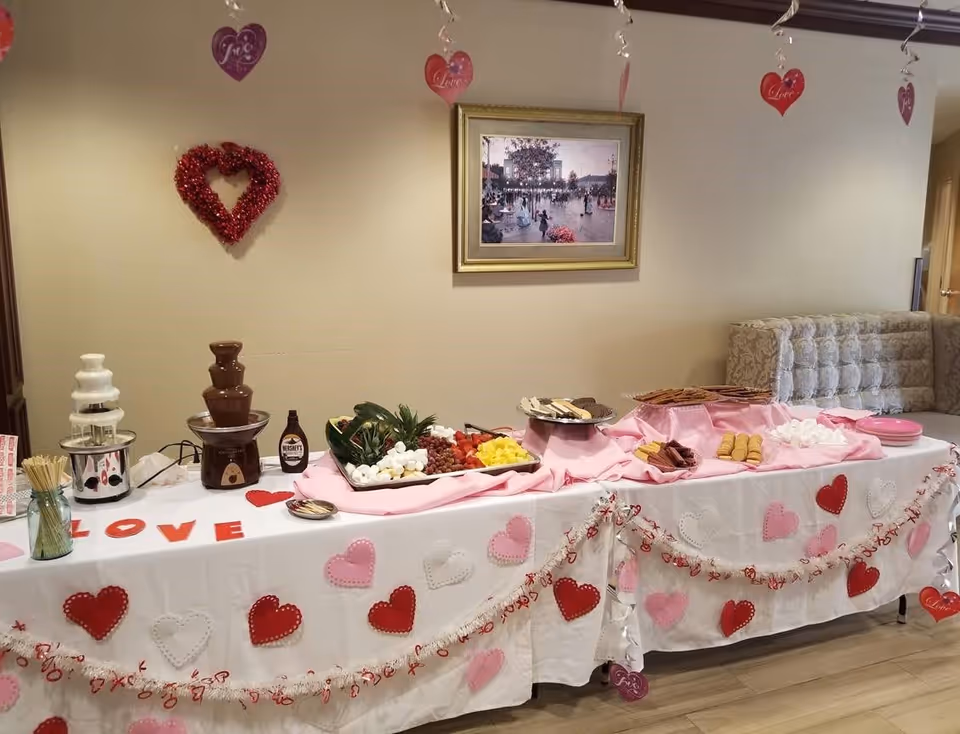 A decorated table set up for a Valentine's Day themed chocolate fondue party with two chocolate fountains, a tray of fruits and marshmallows, plates of cookies and wafers, and pink plates. The table is covered with a white tablecloth adorned with red, pink, and white heart decorations and garlands. Heart-shaped hanging decorations with the words 'Love' and 'Joy' are suspended from the ceiling. A framed painting hangs on the beige wall behind the table, and a cushioned bench is visible to the right.