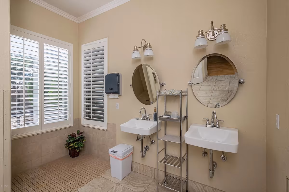 A clean bathroom area with two white sinks mounted on a beige wall, each with a round mirror above and a dual-light fixture. There is a metal shelving unit between the sinks holding toiletries. To the left, there are two windows with white plantation shutters, a small trash bin, and a potted plant on the tiled floor.