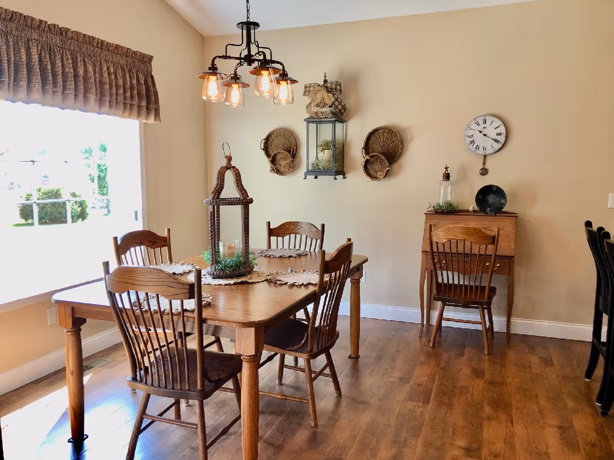 A dining room with a wooden table and four matching wooden chairs. The table is decorated with placemats and a large lantern centerpiece. On the wall, there are three woven baskets and a decorative lantern. A clock and a small wooden desk with a chair are also visible. The room has wooden flooring and a large window with a brown valance letting in natural light.