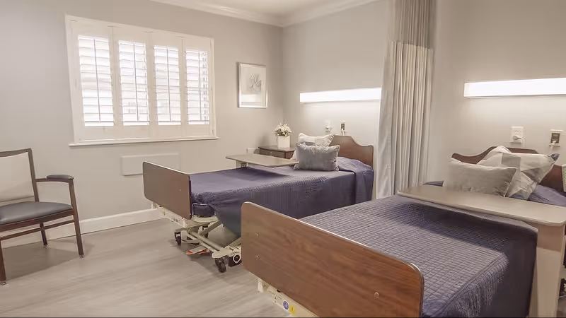 Shared senior care bedroom with two adjustable hospital beds, over-bed tables, a chair, and a window with shutters.