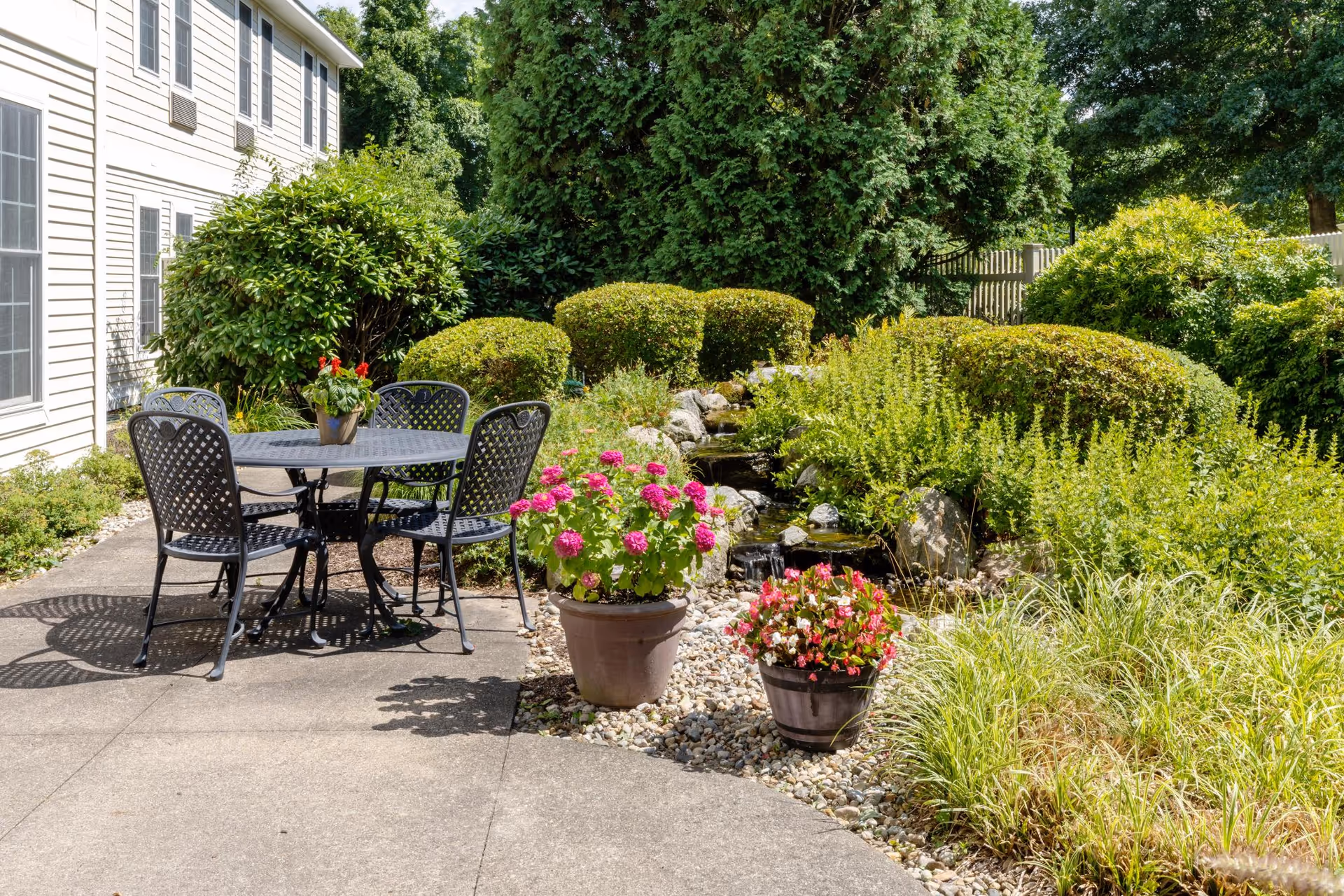 Outdoor patio area with a black metal table and four matching chairs on a concrete surface next to a building. The patio is surrounded by lush green bushes, flowering plants in pots, and a small rock garden with a water feature. Tall trees and a wooden fence are visible in the background.