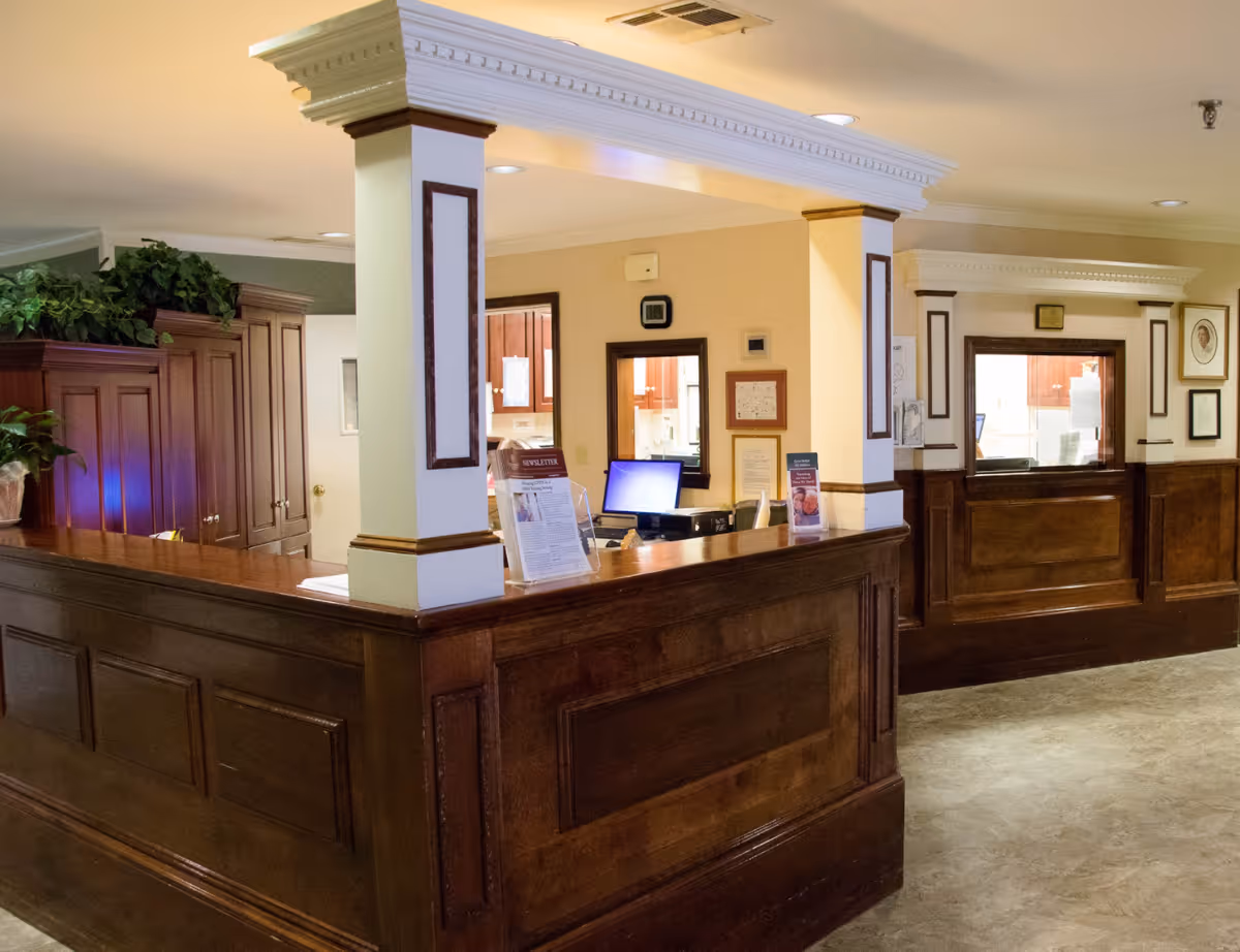 Reception area with a wooden front desk and two service windows in a senior living facility. The desk has informational brochures and a computer monitor. The walls are painted beige and white with decorative molding, and there are plants on top of a wooden cabinet in the background.