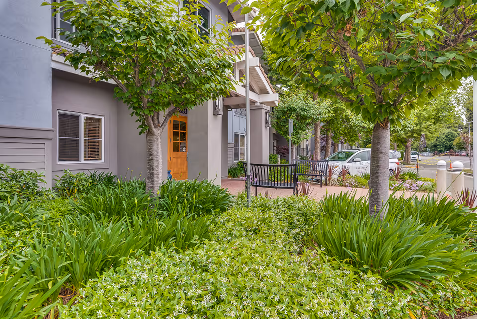 Outdoor view of Pacifica Senior Living Union City showing a landscaped garden with green plants and trees in front of the building entrance. There is a wooden door, windows, a bench, and a parked white car in the background.