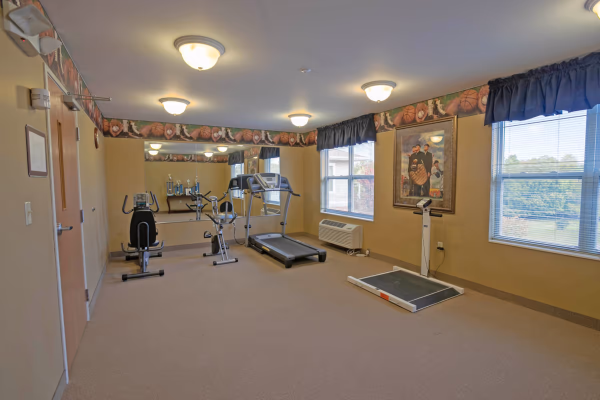A small fitness room with exercise equipment including a treadmill, stationary bike, and a recumbent bike. The room has beige walls with a sports-themed border near the ceiling, featuring images of basketballs, baseballs, and footballs. There are two large windows with dark curtains letting in natural light, a wall-mounted air conditioning unit, a large mirror on one wall, and a painting of three men in vintage baseball uniforms. The floor is carpeted and the room is lit by ceiling-mounted light fixtures.