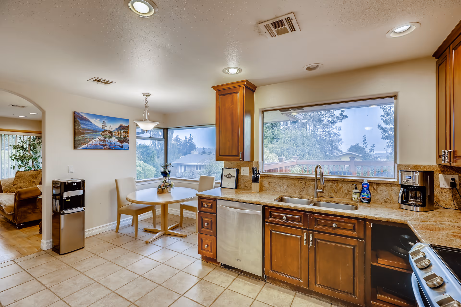 Bright kitchen with wooden cabinets, a stainless dishwasher and sink under a large window, and a small round dining table in a breakfast nook.