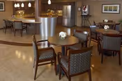Interior view of a senior living facility dining area with round wooden tables and cushioned chairs. The background features a kitchen area with a curved counter, stainless steel refrigerator, and pendant lights hanging from the ceiling. The room has warm lighting and a polished wooden floor.