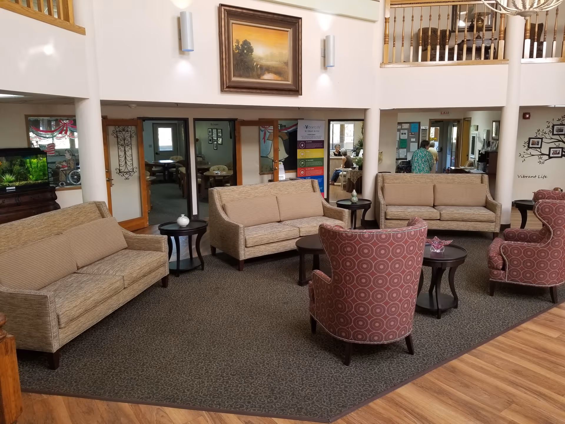 A spacious senior living facility common area with beige sofas, patterned armchairs, and dark wood side tables arranged on a carpeted floor. The room has a high ceiling with a wooden railing balcony above. There is a large framed painting on the wall, a fish tank on a wooden stand, and a colorful informational banner near the entrance to another room. Several people are visible in the background near the entrance and reception area.