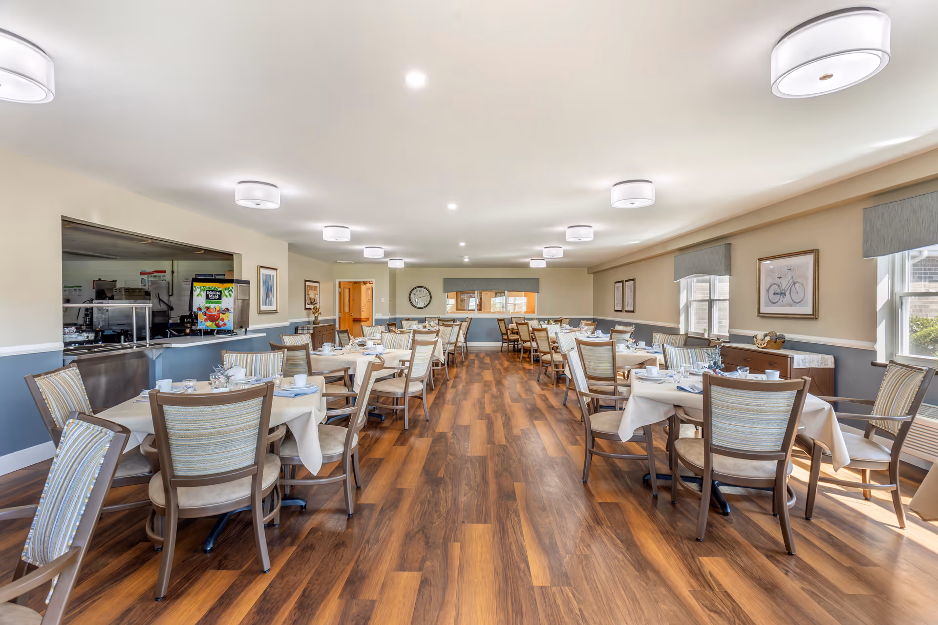 A spacious dining room with multiple tables covered with white tablecloths, each set with plates, glasses, and silverware. The room has wooden flooring, beige walls with a blue-gray wainscot, and several large windows letting in natural light. Ceiling lights are evenly spaced, and there is a serving area with a counter on the left side of the room.