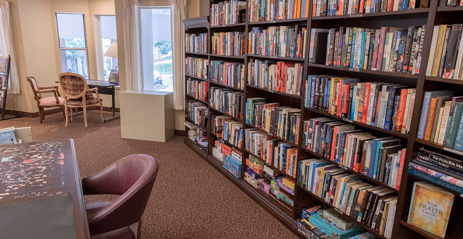 Interior view of a library or reading room in a senior living facility with tall bookshelves filled with books labeled as fiction, a table with a partially completed puzzle, and several chairs near windows letting in natural light.