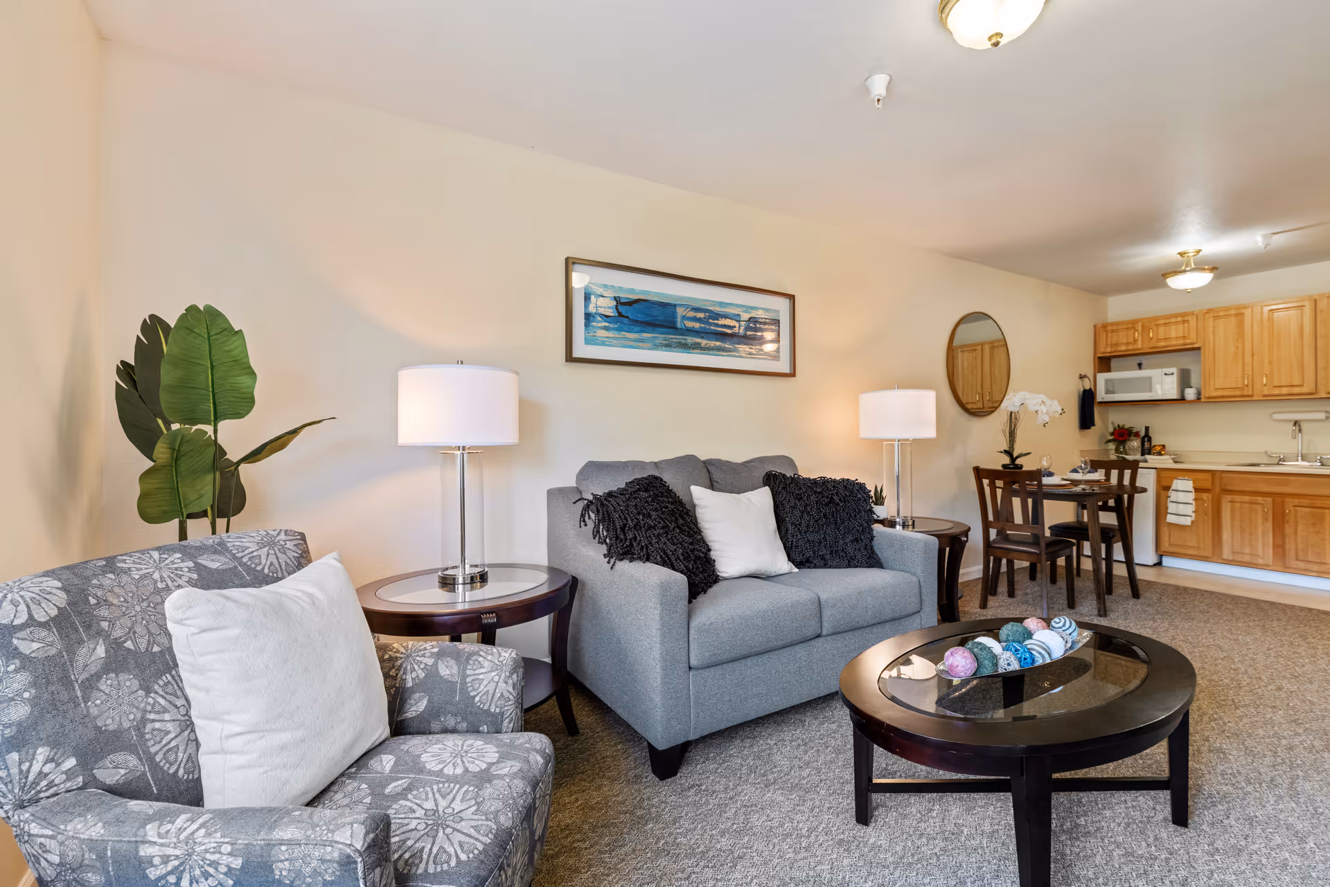 A cozy living room area with a gray loveseat adorned with black and white pillows, a patterned armchair with a white pillow, two round wooden side tables with lamps, a round glass coffee table with decorative balls, a dining table with four chairs, and a kitchen area with wooden cabinets, a microwave, and a sink. The walls are light-colored with a framed artwork and a round mirror.
