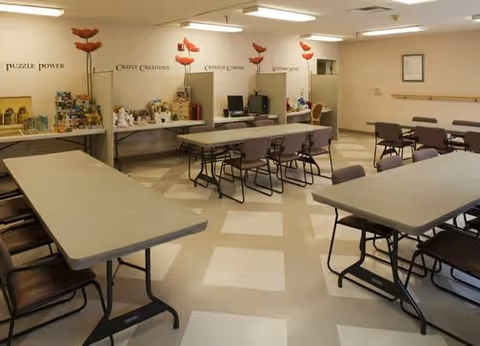 A spacious activity room with several rectangular tables and chairs arranged for group activities. The walls have signs labeled Puzzle Power, Crafty Creations, Cleaning Corner, and Kitchen Spot, indicating different activity stations. The room is well-lit with overhead fluorescent lights and has a tiled floor with a checkered pattern.