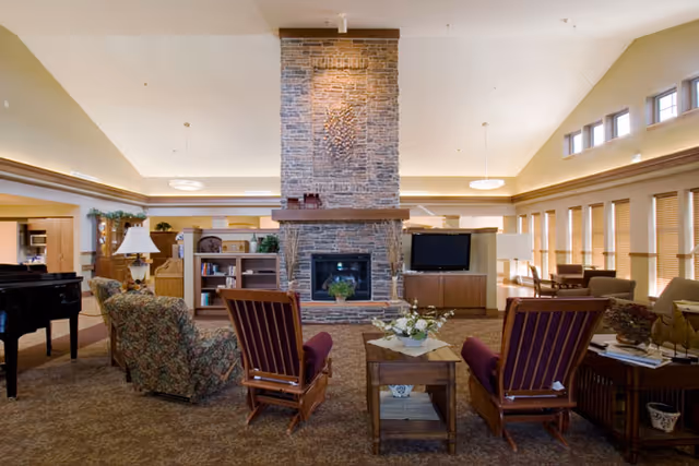 Spacious senior living lounge featuring a large central stone fireplace, seating area with armchairs and sofas, a TV and piano.