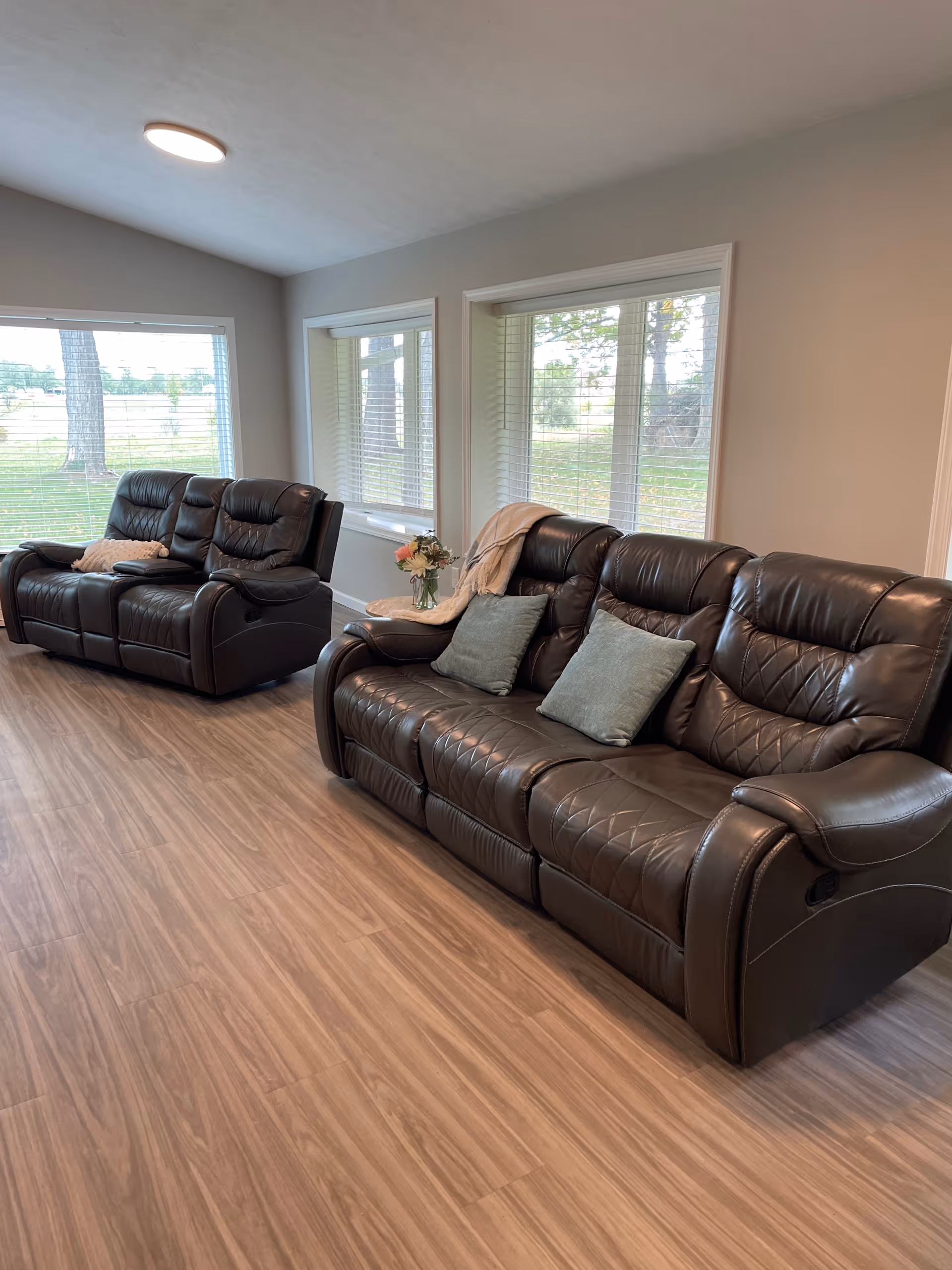 A living room with two dark brown leather reclining sofas, each with cushions. The room has large windows with white blinds letting in natural light, and a small round table with a flower vase and a throw blanket between the sofas. The floor is wood with a light brown finish.