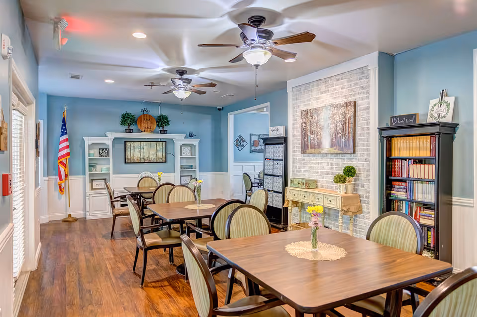A cozy dining room with wooden tables and chairs arranged neatly. The walls are painted blue with white wainscoting, decorated with framed artwork and shelves holding books and plants. Ceiling fans with lights hang from the ceiling, and small vases with flowers are placed on the tables.