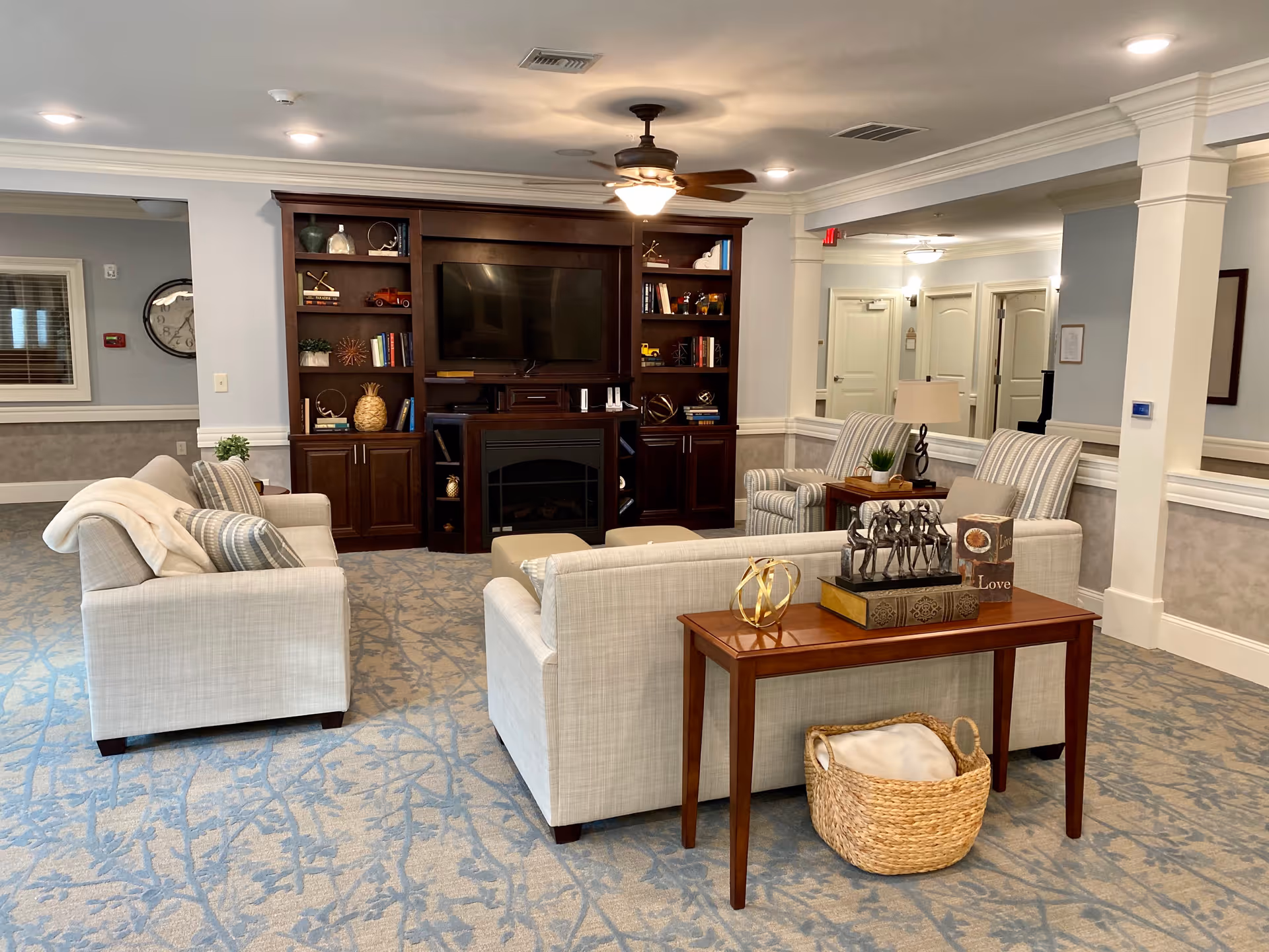 A cozy living room area in a senior living facility featuring two beige sofas, two striped armchairs, a wooden entertainment center with a TV and decorative items, a ceiling fan with light, and a wooden console table with decorative objects and a woven basket underneath. The room has soft blue patterned carpet and neutral-colored walls with white trim.