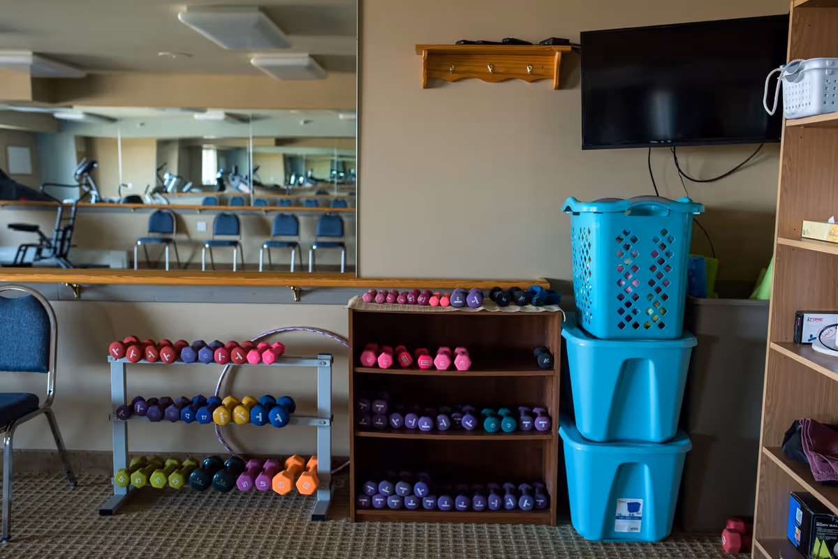 Exercise corner with shelves of colorful dumbbells, stacked turquoise storage bins, a wall-mounted TV, and a large mirror reflecting chairs and equipment.