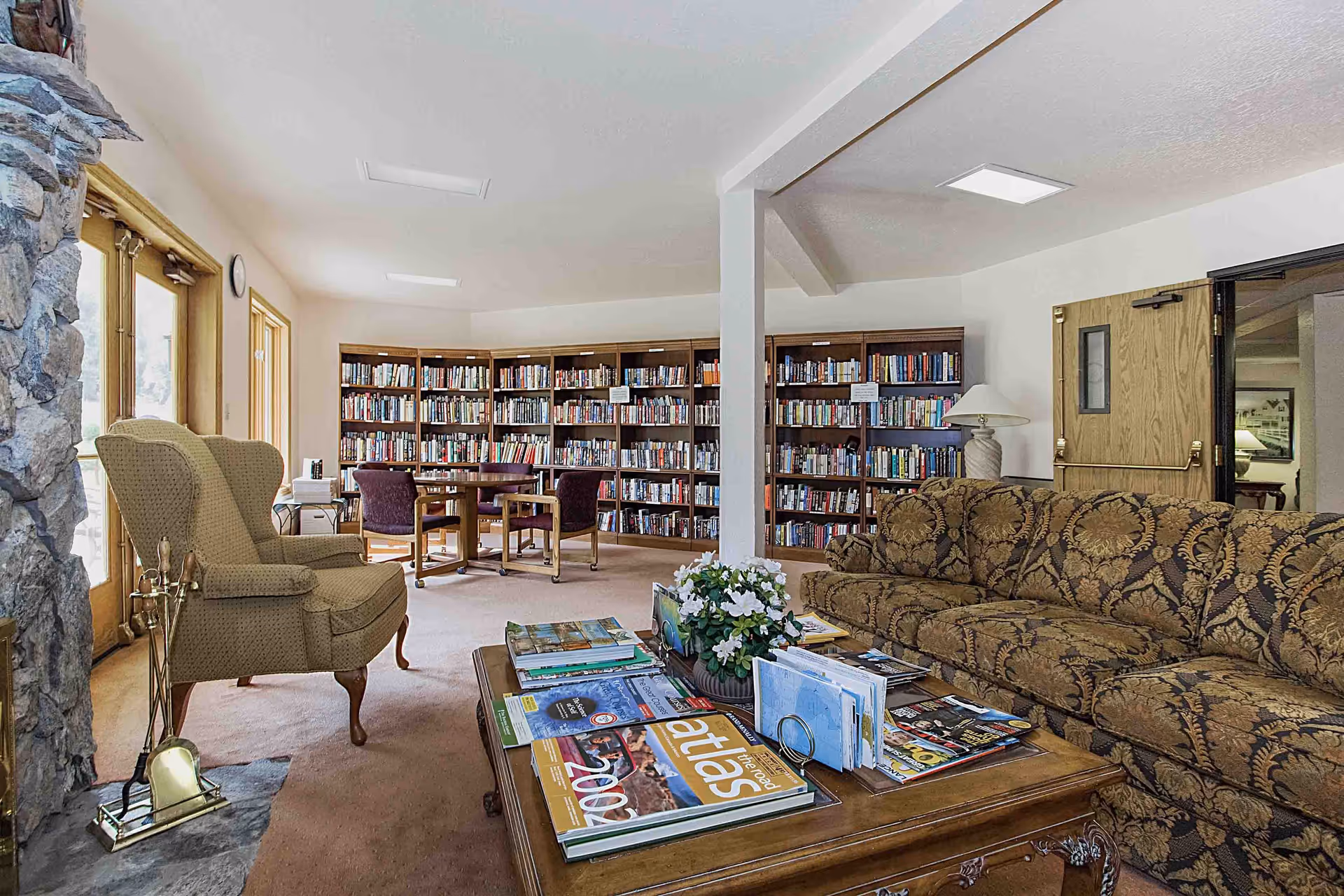A cozy reading room in a senior living facility featuring a patterned sofa, an armchair, a wooden coffee table with magazines and books, a stone fireplace, and a wall lined with bookshelves filled with books. There is also a round table with chairs near the bookshelves and large windows letting in natural light.