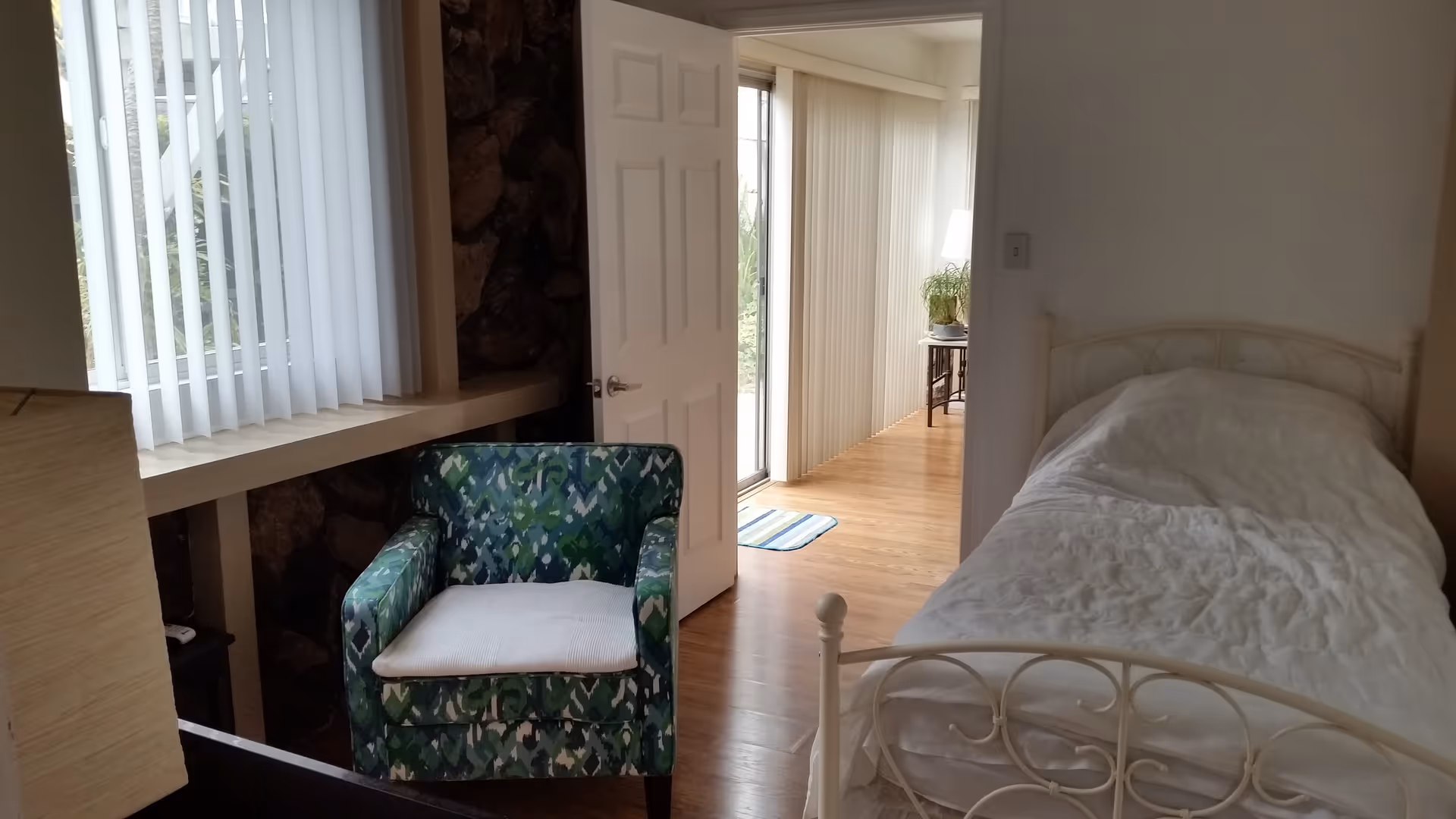 A small bedroom with a single white metal bed frame and a white comforter. Next to the bed is a green patterned armchair with a white cushion. The room has a window with vertical blinds and a door that opens to a hallway with wooden flooring and a small table with a plant and lamp.