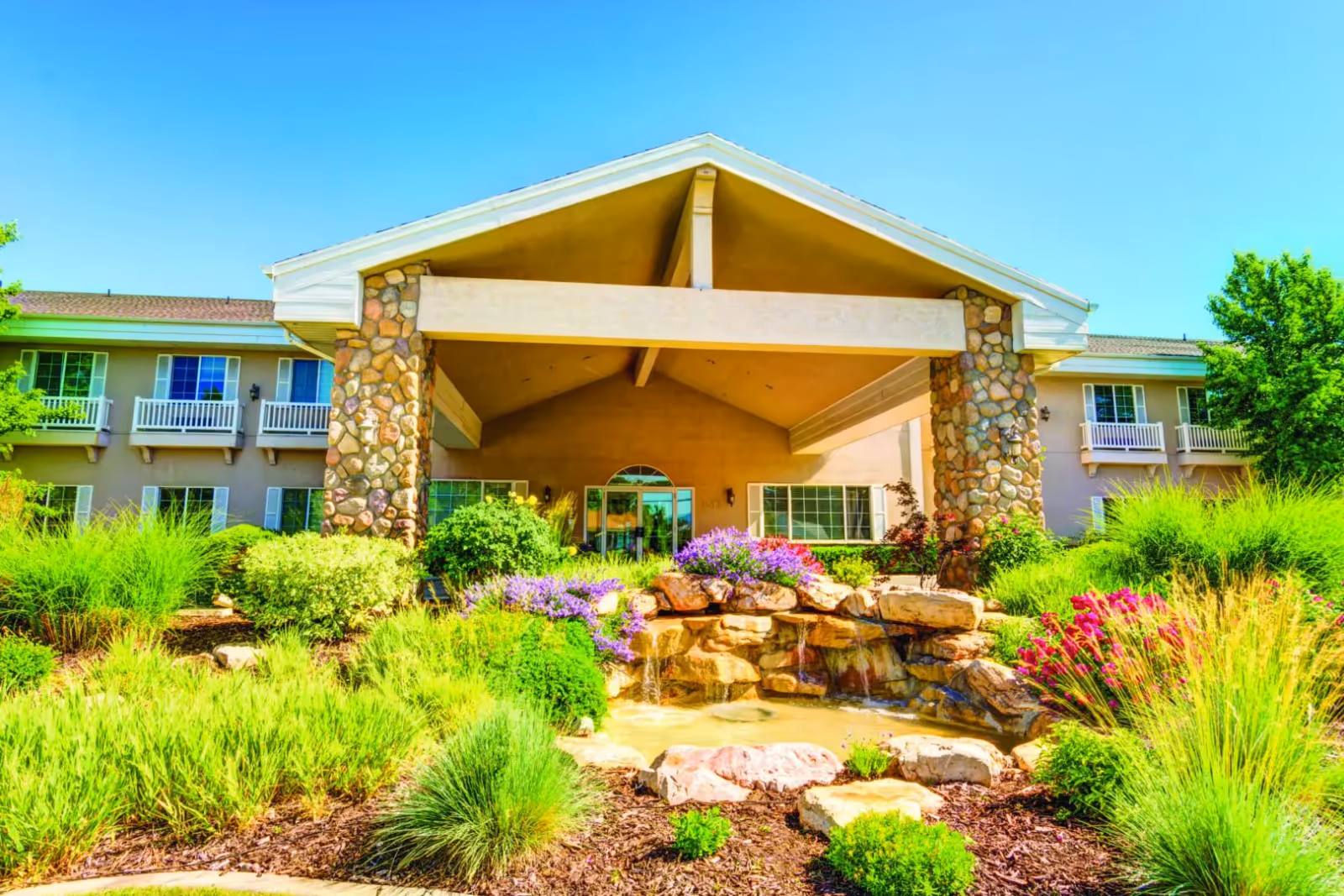 Front exterior view of The Wentworth at East Millcreek facility featuring a covered entrance supported by stone pillars, surrounded by lush green landscaping with various bushes, flowering plants, and a small rock water feature under a clear blue sky.