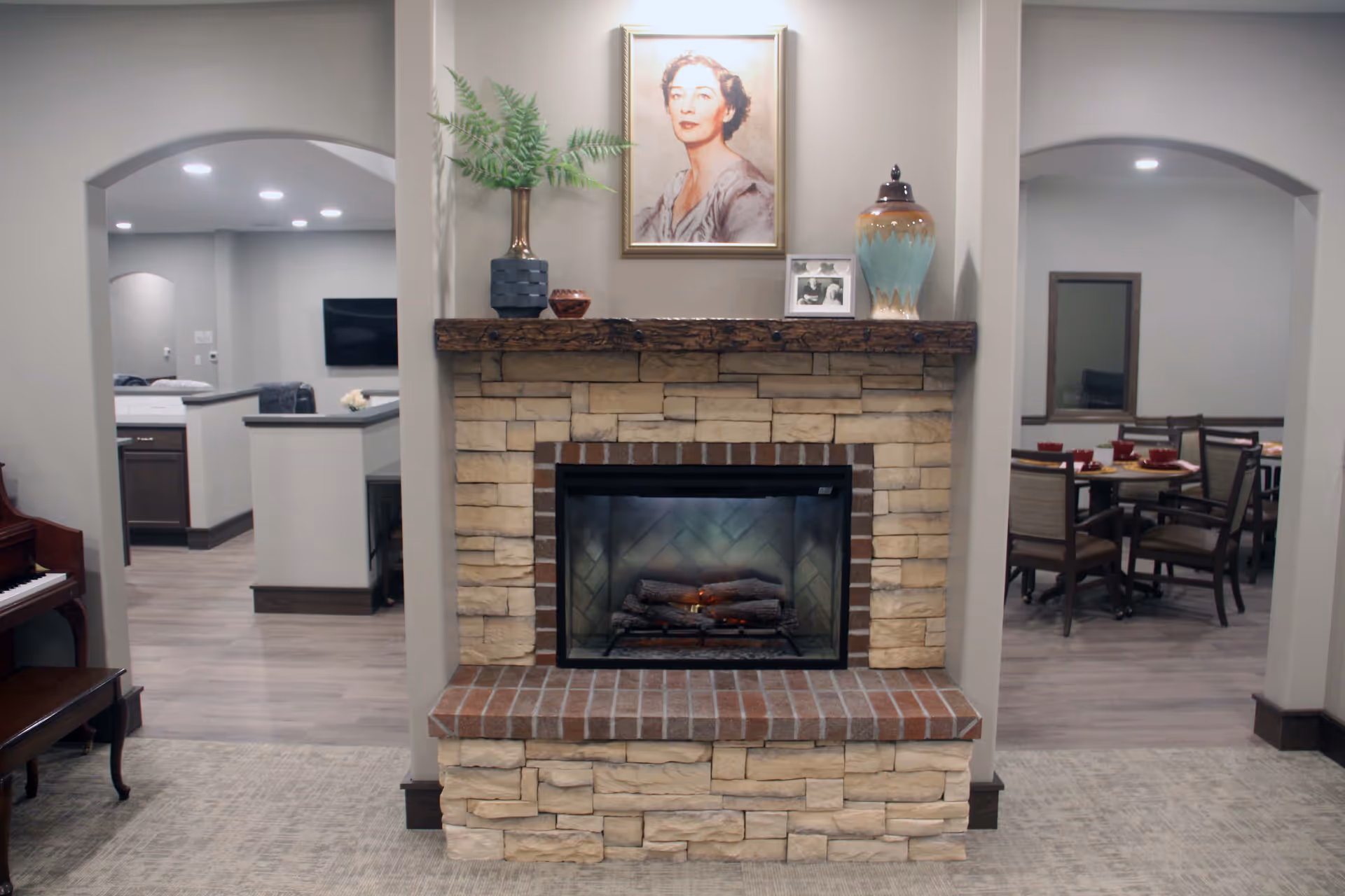 A cozy interior space featuring a stone and brick fireplace with a wooden mantel. On the mantel are decorative items including a potted plant, a framed photograph, a ceramic vase, and a portrait of a woman hanging above. To the left, there is a piano, and through an archway, a kitchen area is visible. To the right, another archway leads to a dining area with tables and chairs set for a meal.