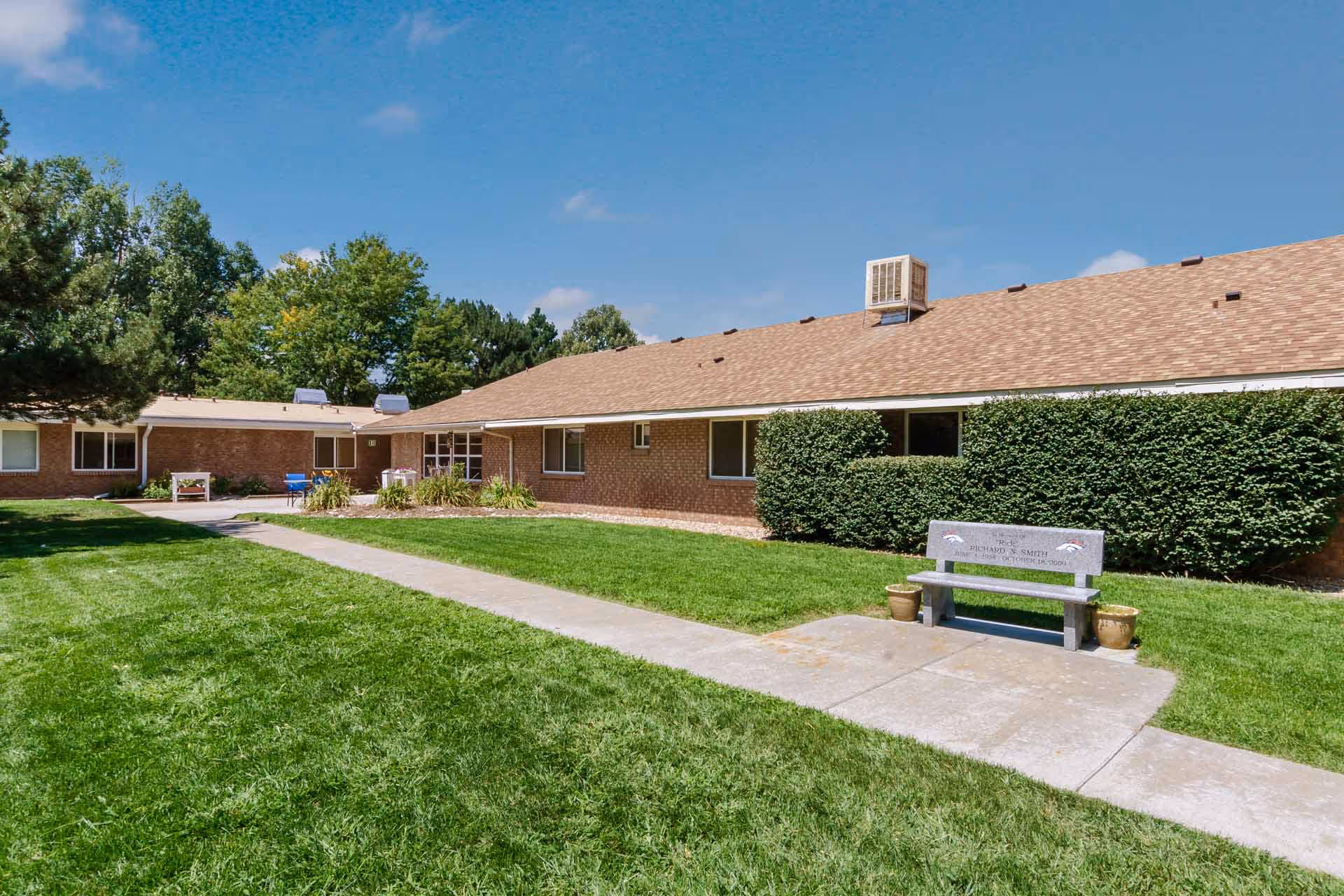 Single-story brick senior living building with a manicured lawn, sidewalk, and a commemorative bench in the courtyard.