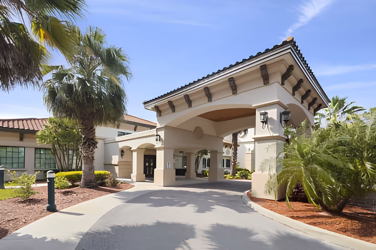 Entrance of Discovery Commons South Biscayne facility featuring a covered driveway with beige pillars and a tiled roof, surrounded by palm trees and landscaped greenery under a clear blue sky.