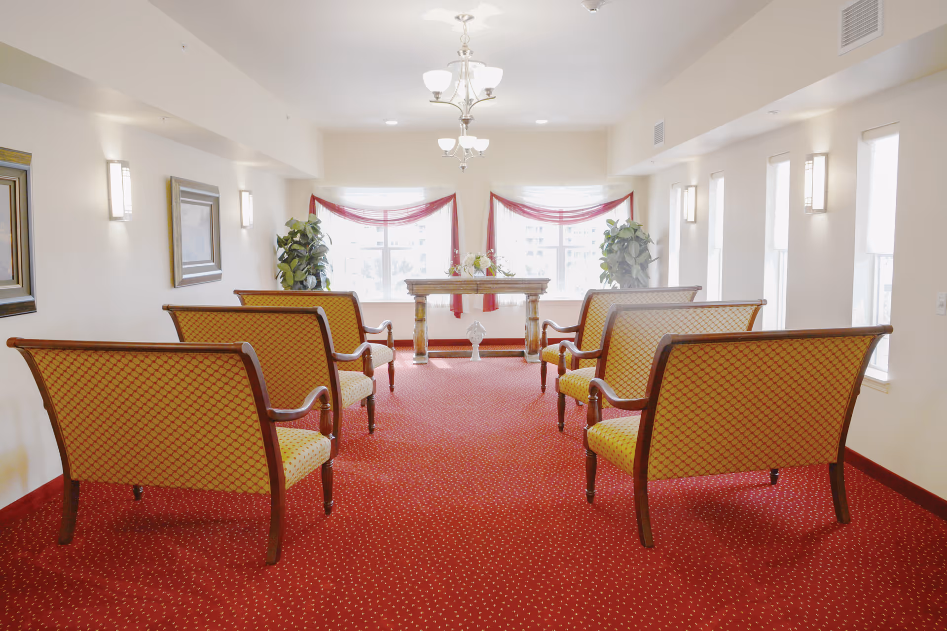 Bright communal sitting room with upholstered benches arranged facing a table by large windows and red carpet.
